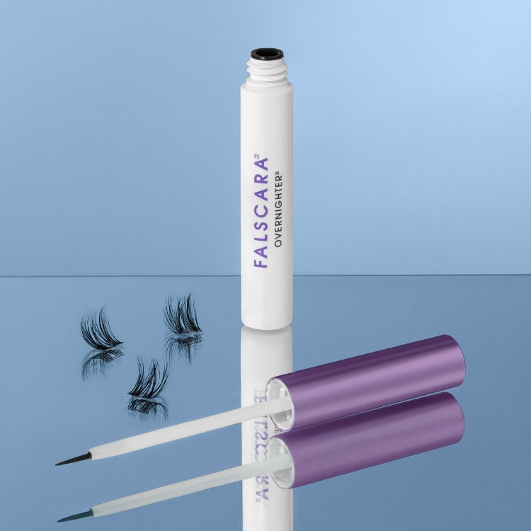 Falscara - Overnighter, witte tube met paarse dop en applicator, naast zwarte wimpers op een spiegelachtige ondergrond.