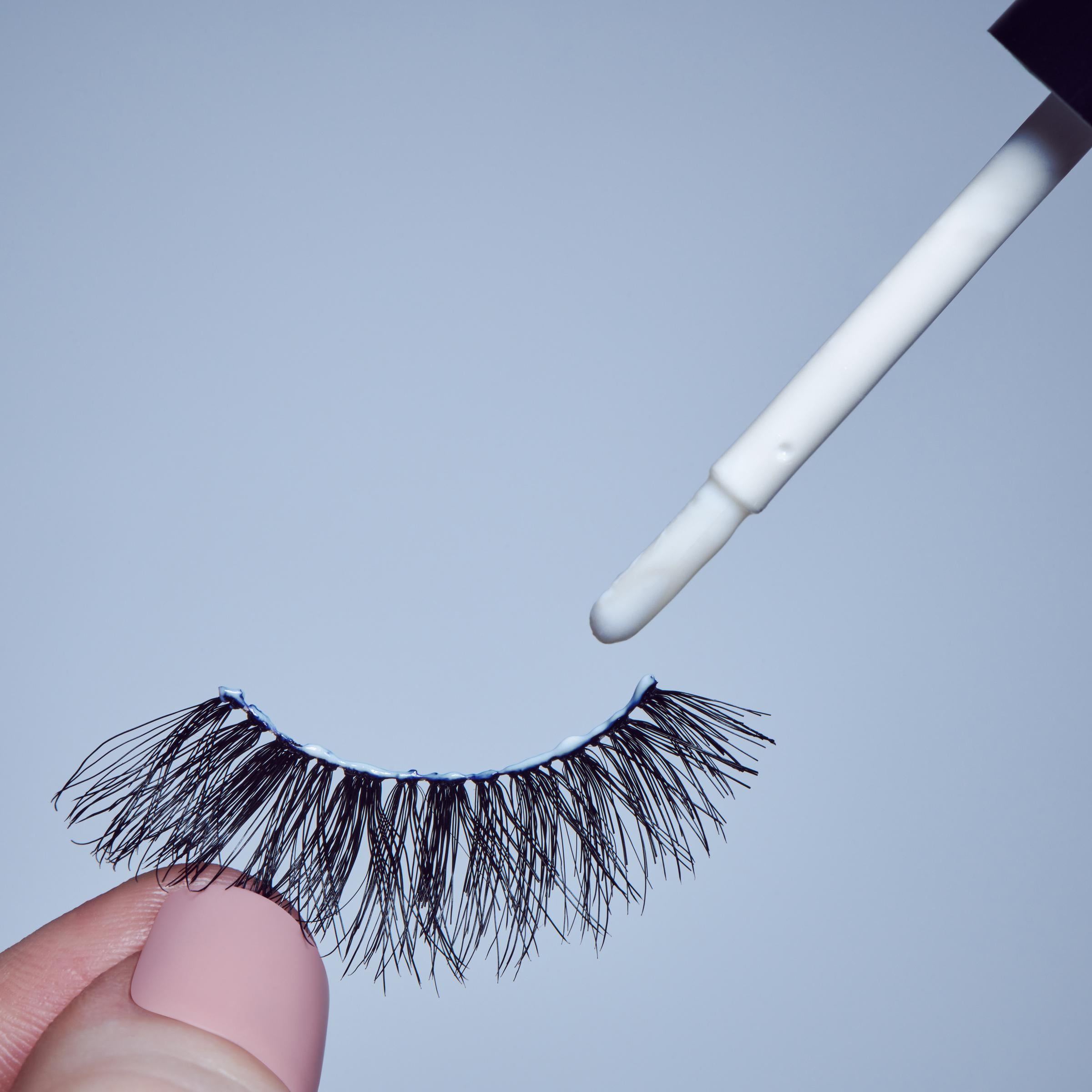 Wimperlijm Lash Glue - Clear in een witte verpakking met een applicator, naast een paar zwarte wimpers.