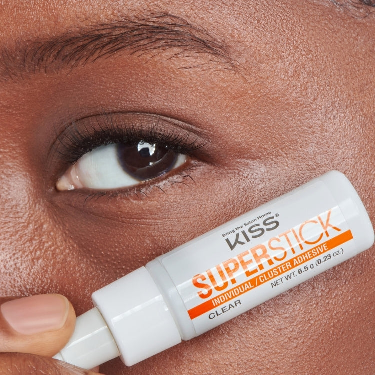 Superstick Cluster Lash Adhesive | Clear in een witte tube met oranje en zwarte tekst, aangebracht op een hand.