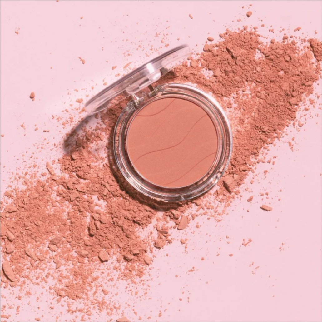 Soft Glow Blusher in een transparante verpakking met een warme perzikkleur, omringd door losse poederdeeltjes.